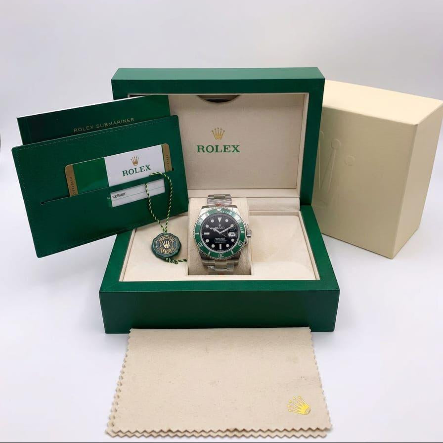 Rolex watch presented in an open green Rolex gift box with a cream insert, alongside a green booklet, a cream outer box, and a cream dust bag. The watch has a silver-toned metal case, a black dial, and a green bezel.
