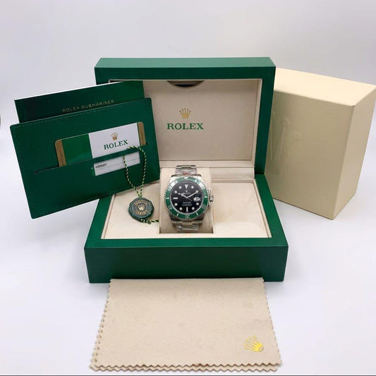 Rolex watch presented in an open green Rolex gift box with a cream insert, alongside a green booklet, a cream outer box, and a cream dust bag. The watch has a silver-toned metal case, a black dial, and a green bezel.
